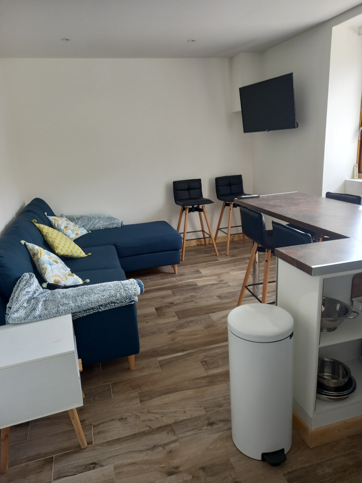 A modern living area features a dark blue sectional sofa adorned with patterned cushions, complemented by a high-top dining table with four black stools. A flat-screen television is mounted on the wall, and a white bin is positioned nearby, adding to the room's functionality.