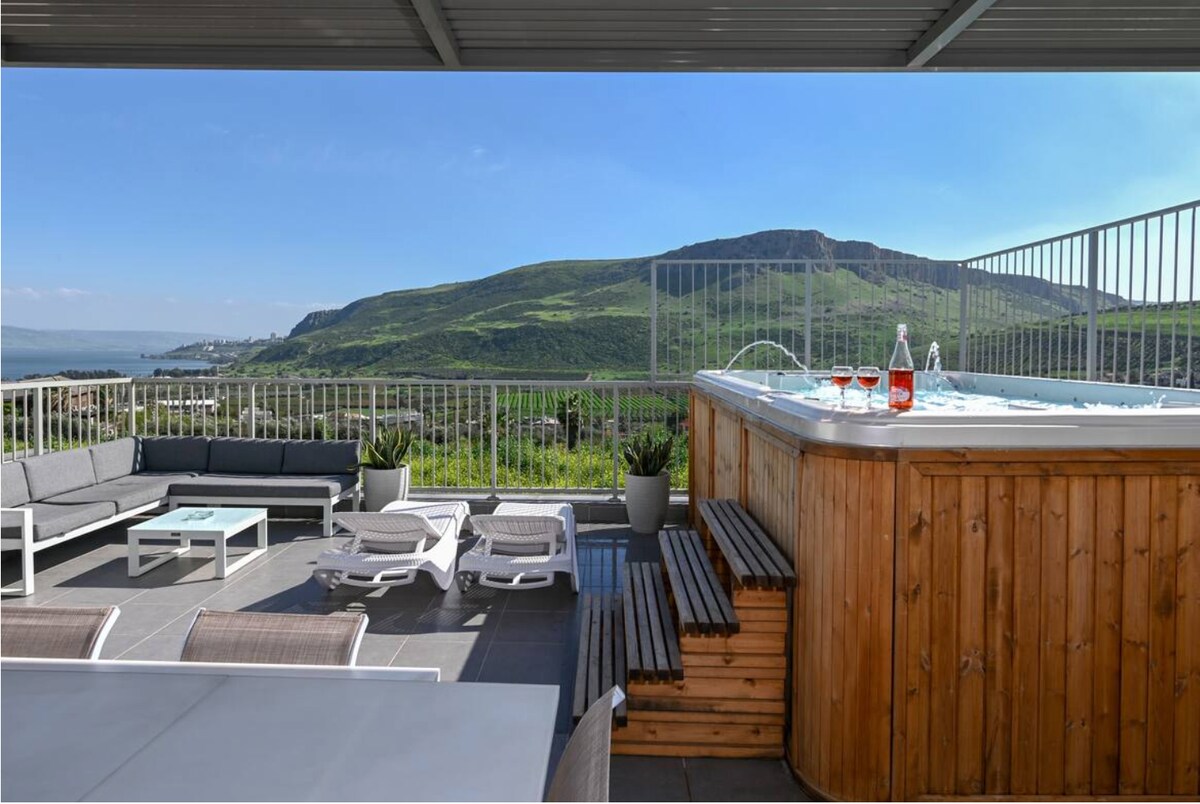 A spacious balcony features a wooden hot tub, surrounded by seating areas and sunbeds. The view highlights lush hills and the Sea of Galilee in the distance, with clear skies and sunlight illuminating the outdoor space.