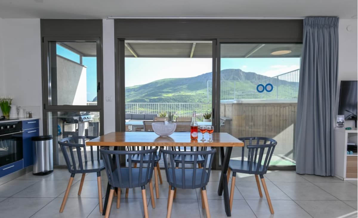 A dining area is visible with a wooden table surrounded by six blue chairs. Large sliding glass doors frame a mountain view, allowing natural light to fill the space. A bowl with greenery and two bottles sits on the table, enhancing the inviting environment.