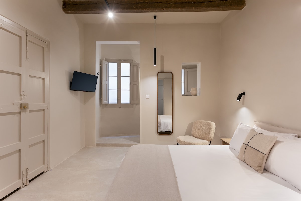 A clean, minimal bedroom features a spacious double bed with white linens. A smart TV is mounted on the wall opposite. A mirror and a light-colored chair add functionality. Natural light enters from windows, creating an airy feel, with exposed wooden beams highlighting the ceiling.