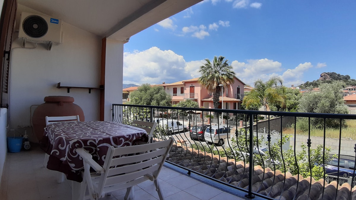 A balcony features a table covered with a patterned cloth and two white chairs. A view of the surrounding greenery and nearby buildings is visible, with a blue sky and scattered clouds providing a serene backdrop.