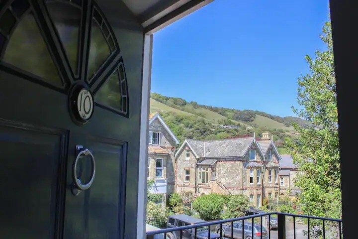 A welcoming view from the entrance captures the vibrant greenery of rolling hills, along with charming neighboring houses. The clear blue sky provides a bright backdrop, creating an inviting atmosphere for guests arriving at the property.