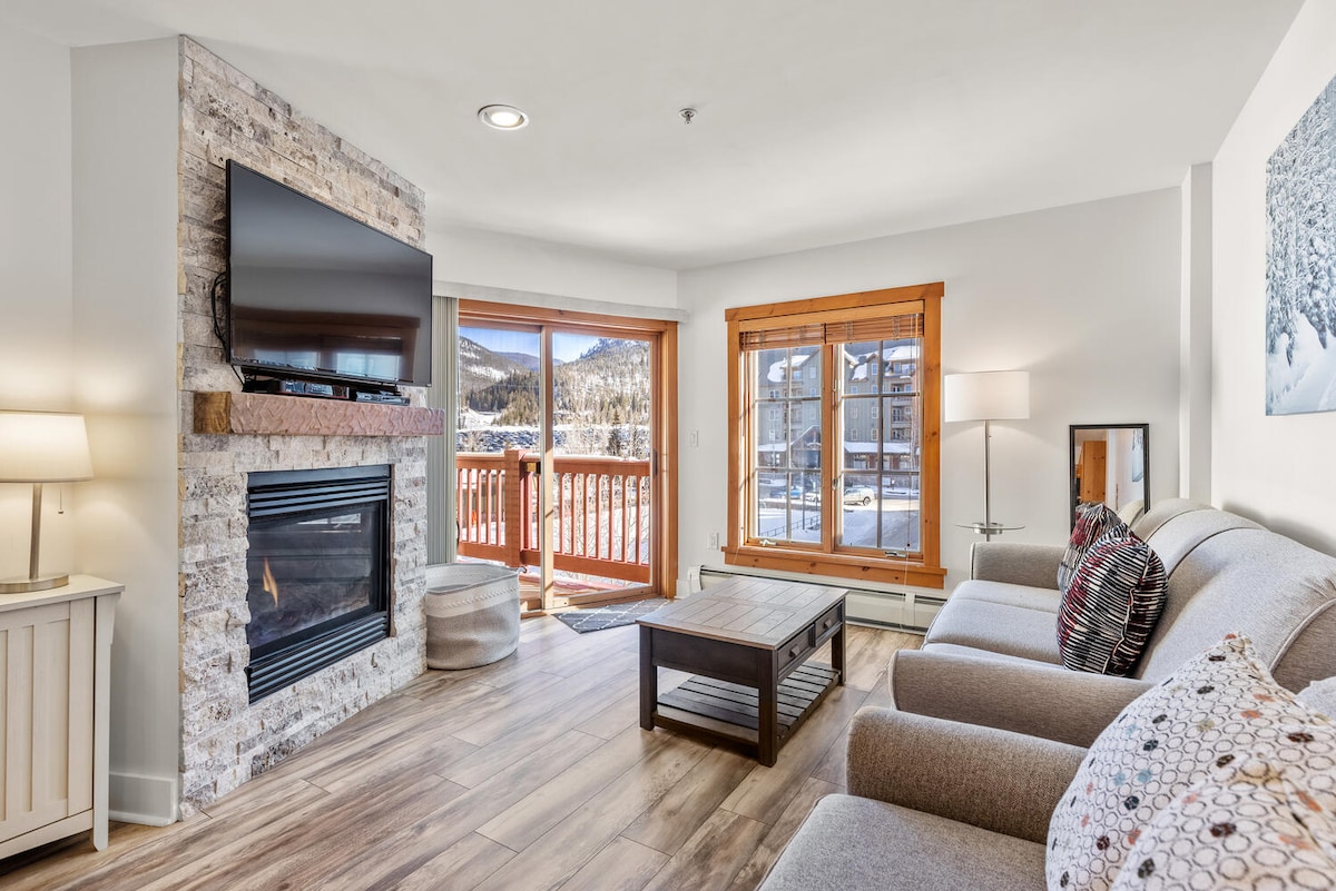 A cozy living room features a soft sofa and a coffee table in front of a stone-accented gas fireplace. Large windows provide mountain views and allow natural light to fill the space. A sliding door leads to a balcony, enhancing the outdoor connection.