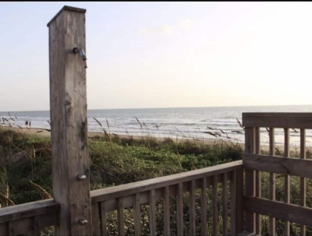 A wooden shower fixture is positioned near a pathway leading to the beach. Dune grasses and shrubs frame the scene, while the ocean stretches into the horizon under a soft sky. People can be seen walking along the shoreline in the distance.
