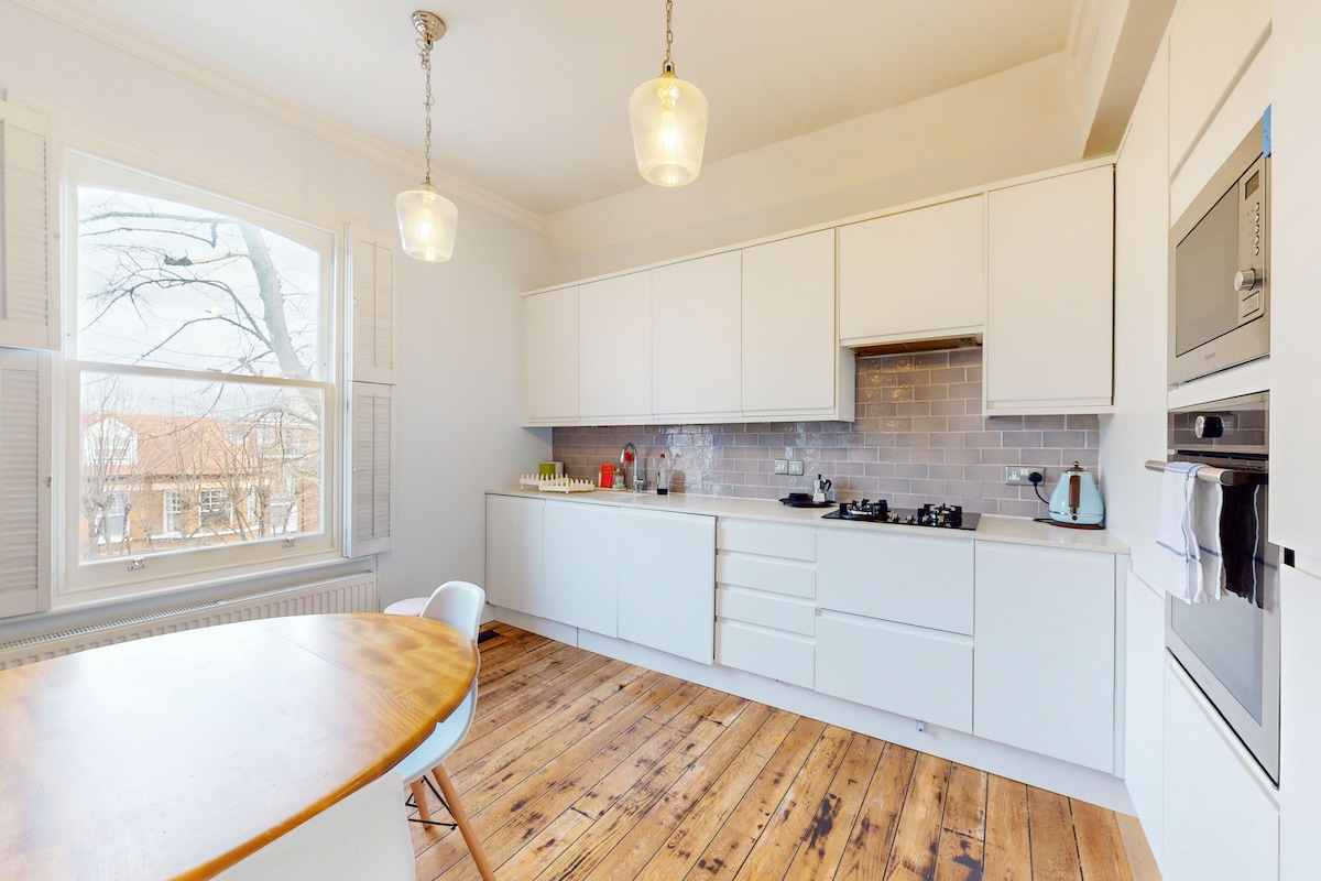 A modern kitchen is highlighted by sleek white cabinetry and a built-in gas stove. Natural light floods through a large window, illuminating the wooden floor. A round dining table with white chairs complements the space, creating a practical yet inviting atmosphere.