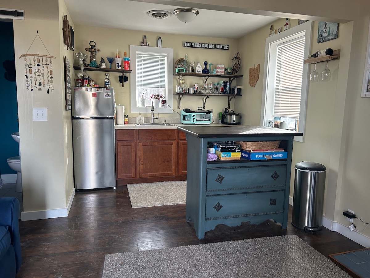 A functional kitchen is showcased, featuring wooden cabinets and a silver refrigerator. Open shelving displays various kitchen items. A spacious island with a colorful finish forms the central point. Natural light enters through windows, illuminating the area and creating a warm atmosphere.