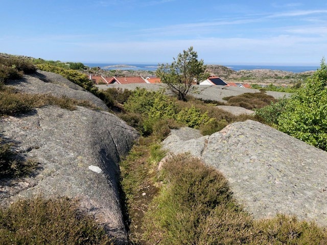 A rocky landscape is presented, showcasing a pathway winding between boulders and patches of greenery. In the background, the ocean becomes visible under a clear blue sky, with distant land formations adding depth to the serene view.