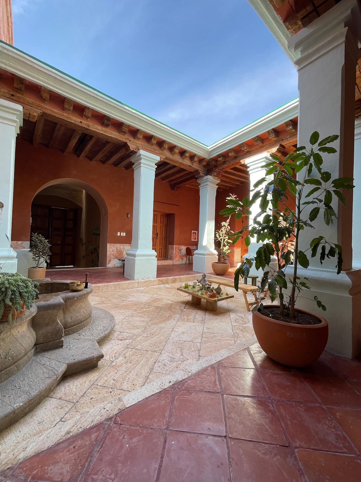 A central patio is framed by tall columns and terracotta walls, featuring a stone fountain surrounded by lush plants in decorative pots. The floor is made of patterned tiles, and there are two small seating areas that invite relaxation and social interaction.