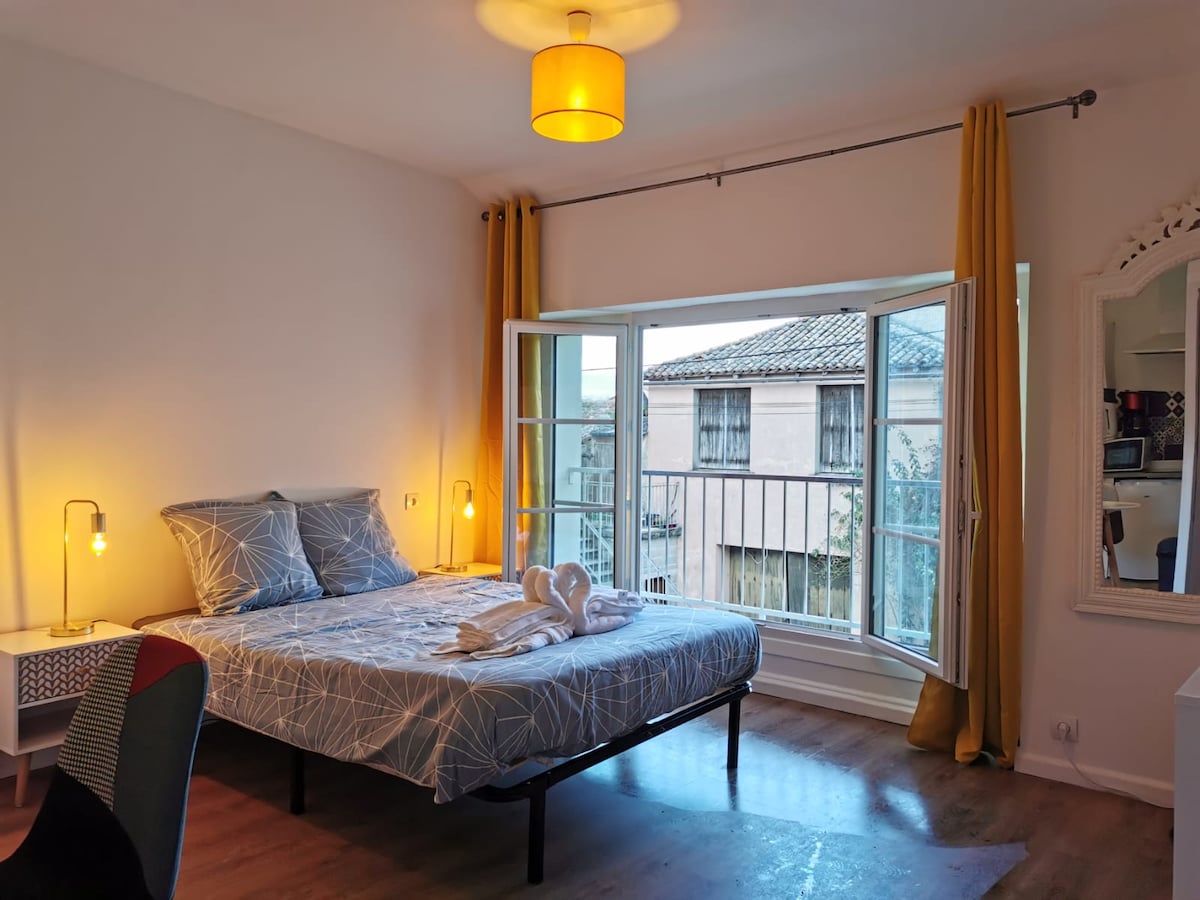A bright bedroom features a spacious bed dressed in light linens with a geometric pattern. Large windows with yellow curtains provide natural light and a view, while a bedside table with a lamp adds functionality. The floor is finished in warm wood.