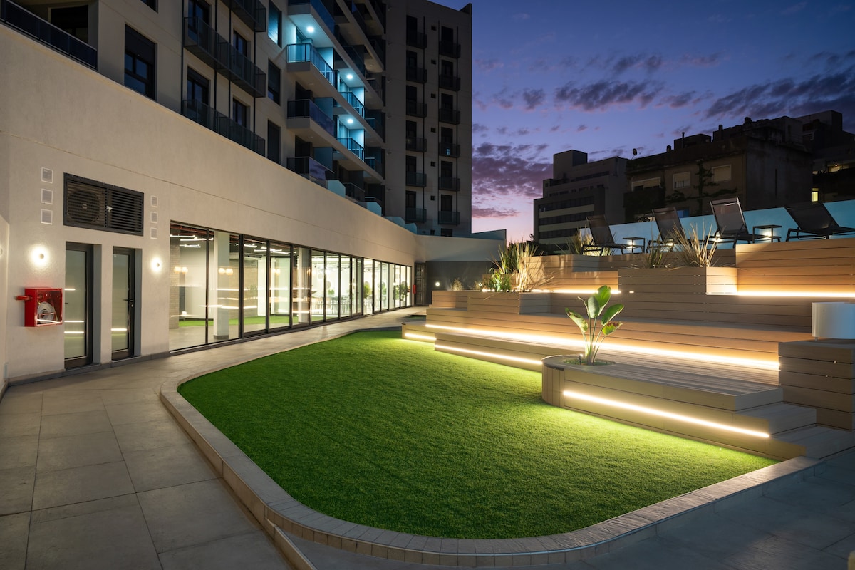 A modern outdoor area is illuminated by soft lighting, featuring a green lawn bordered by sleek wooden seating. The backdrop showcases the building's contemporary architecture under a twilight sky, highlighting large glass windows that reflect the evening ambiance.