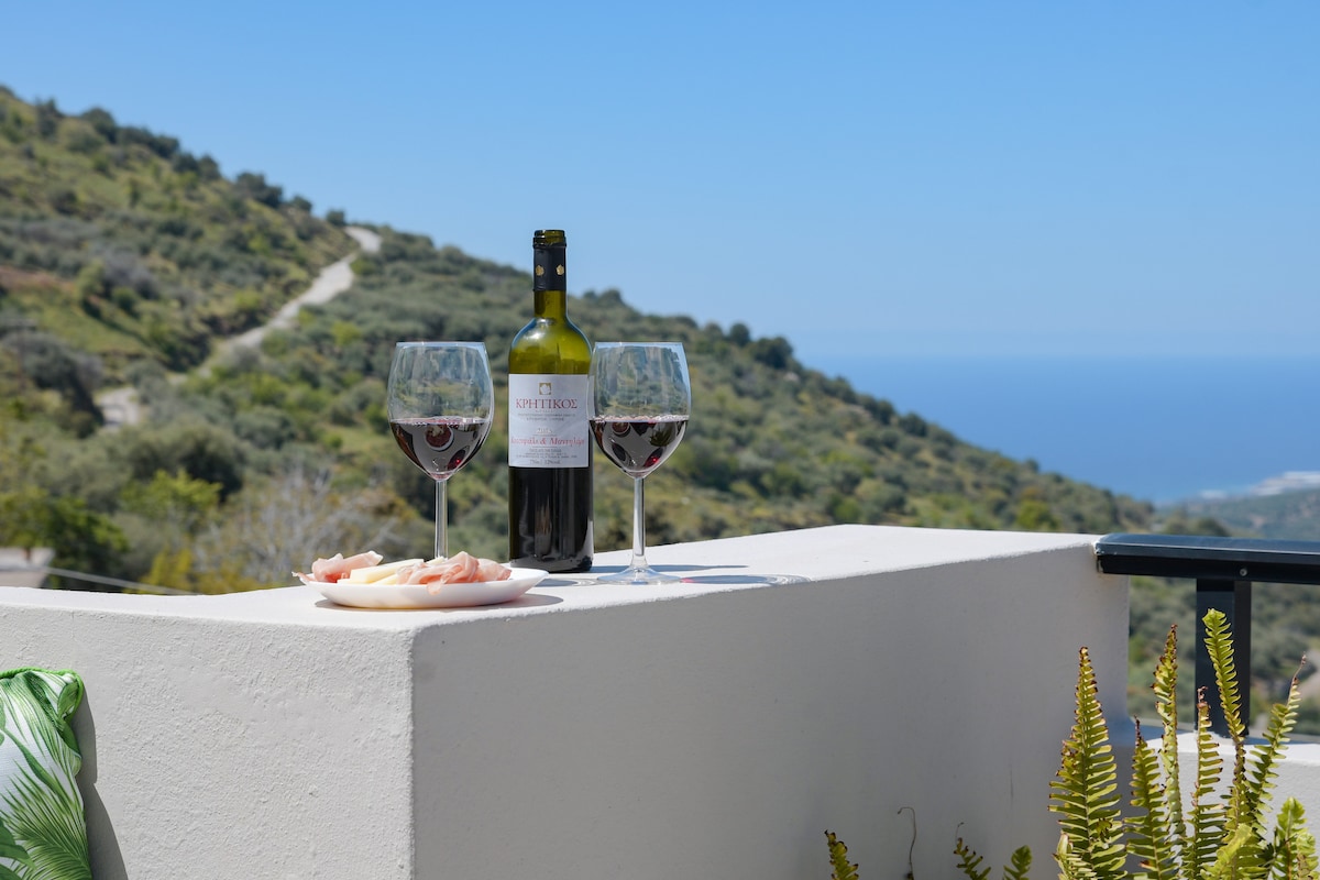 A bottle of red wine is positioned between two glasses, both filled to the brim. A plate with sliced cheese and charcuterie sits nearby. In the background, the hillside and the deep blue sea create a serene view, highlighting the tranquil setting.