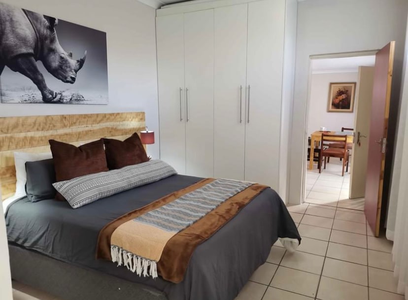 A cozy sleeping area features a large bed with decorative pillows and a warm throw blanket. A striking image of a rhinoceros is displayed above the headboard. The space includes built-in wardrobes and a doorway leading to a dining area with visible furniture.