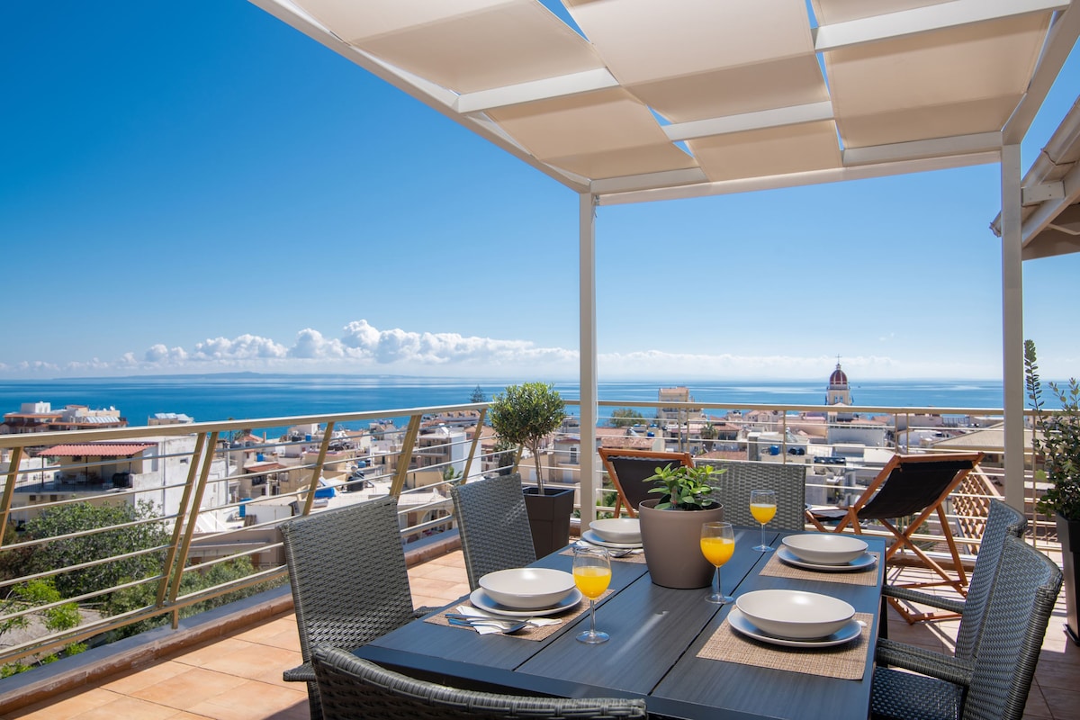 A spacious terrace is furnished with a dining table set for four, complemented by refreshing orange drinks. Comfortable lounge chairs provide a relaxing spot. The expansive view offers a blend of cityscape and sea, under a clear blue sky with scattered clouds.