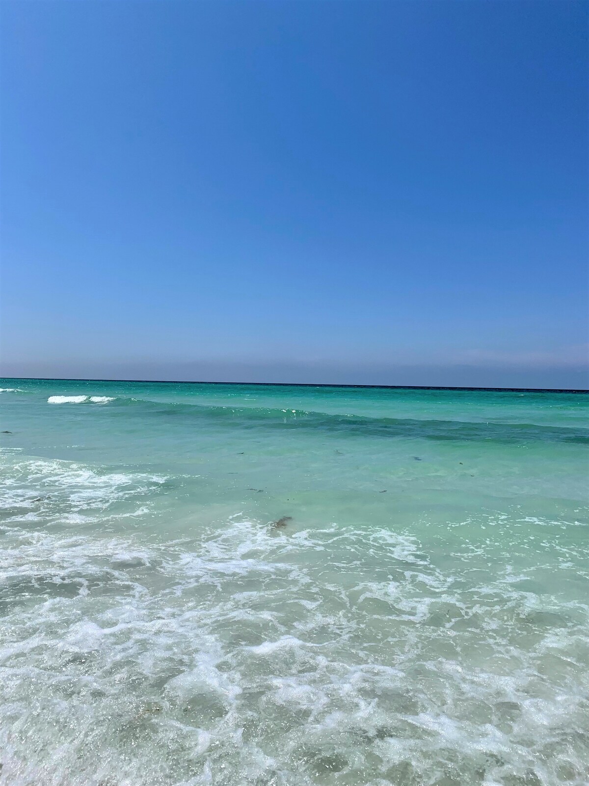 The image captures a serene beach scene with gentle waves rolling onto soft, white sand. The clear turquoise waters extend to the horizon, blending with a bright blue sky. This tranquil environment offers an inviting atmosphere for relaxation.