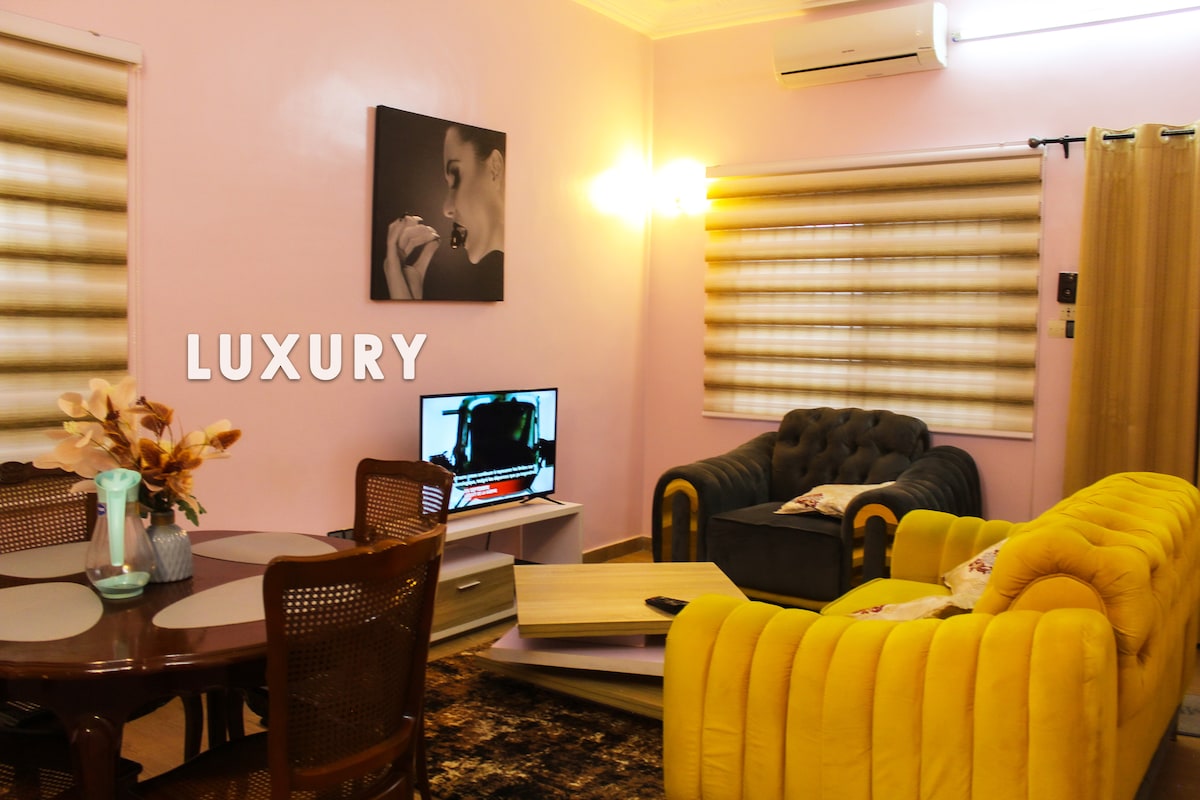 The living area features a combination of yellow and dark blue sofas, accompanied by a wooden dining table with woven chairs. A television is positioned in the corner, while soft lighting enhances the serene atmosphere within the pink-walled space.