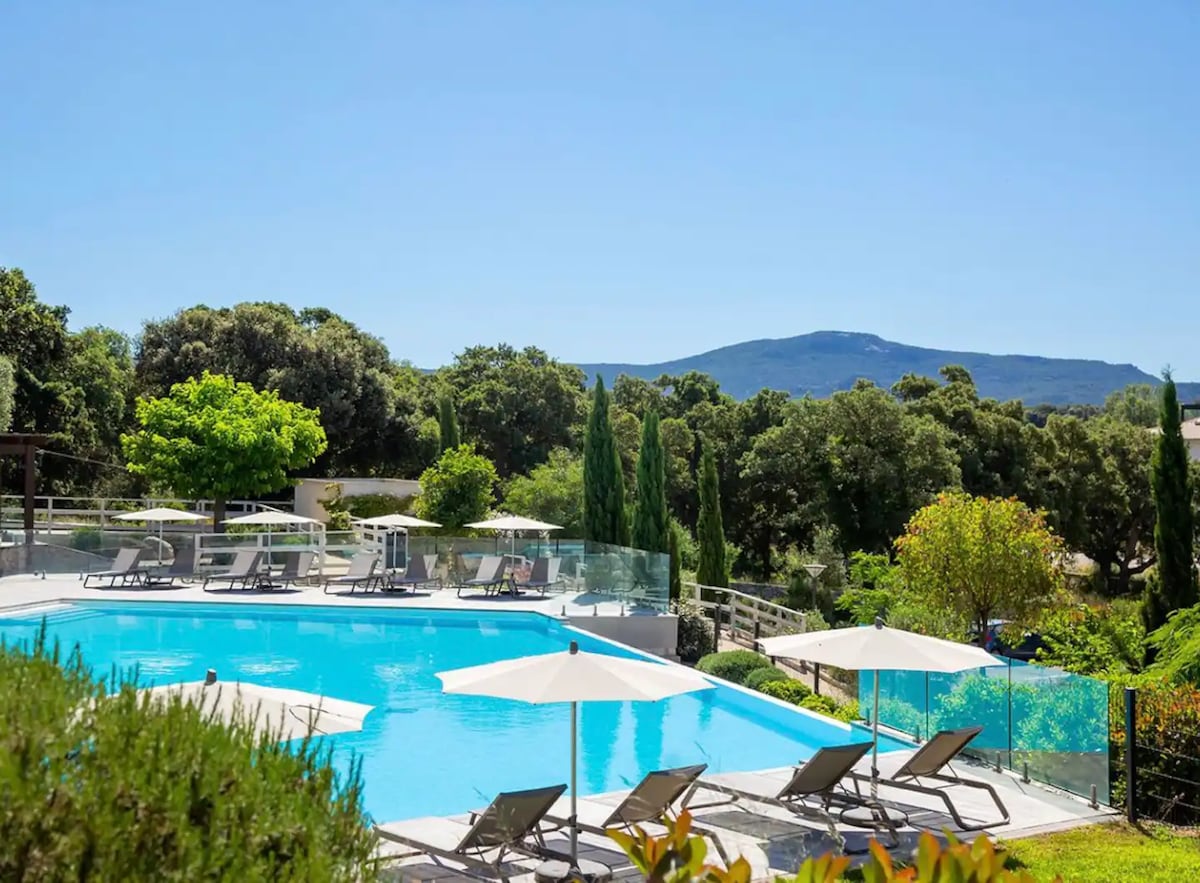 A spacious outdoor pool is set within a lush landscape, featuring sun loungers under umbrellas. Verdant trees and a mountain view create a serene backdrop, while a clear blue sky enhances the peaceful atmosphere of the area.