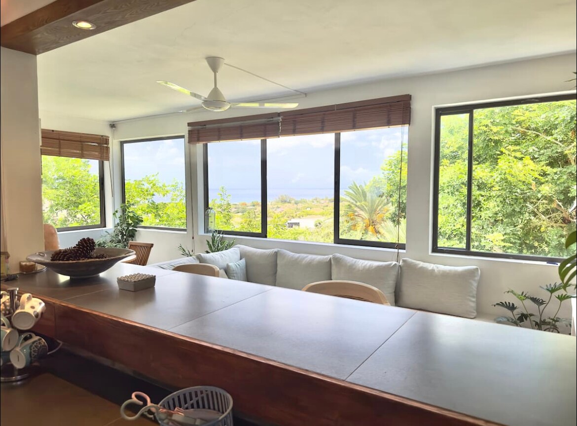 A spacious living area features large windows offering views of greenery and the sea. A cozy seating arrangement is present, adorned with soft pillows. A wooden countertop can be seen in the foreground, complemented by decorative items and kitchen essentials.
