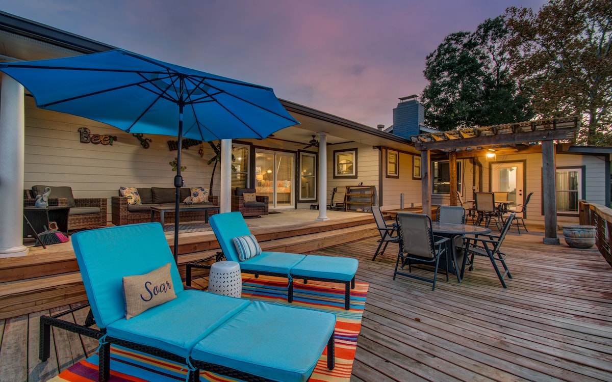 An outdoor deck is presented with lounge chairs featuring blue cushions and a large blue umbrella. The space is equipped with a dining area and various seating arrangements, surrounded by a serene setting at dusk, with warm light glowing from inside the house.