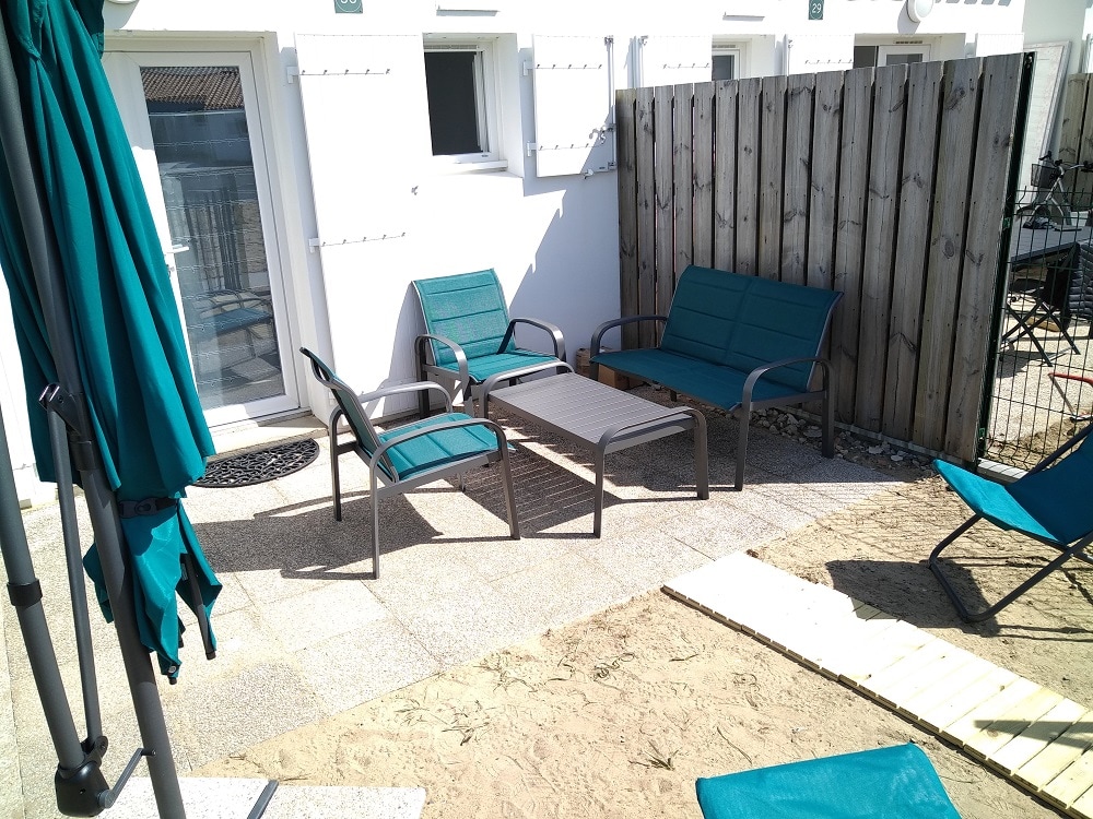 An outdoor seating area is featured, furnished with two lounge chairs and a small table. A shaded umbrella stands nearby. The space is surrounded by a wooden fence, providing privacy. The ground is composed of sand and stone tiles, enhancing the relaxed atmosphere.