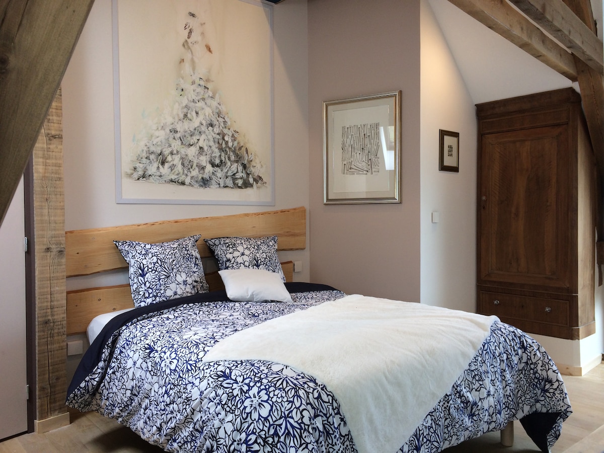 A spacious bedroom features a wooden bed frame topped with a floral patterned duvet. Artistic wall decor adorns the walls, complemented by a brown wooden wardrobe. Natural light filters in, creating a serene and calming ambiance.