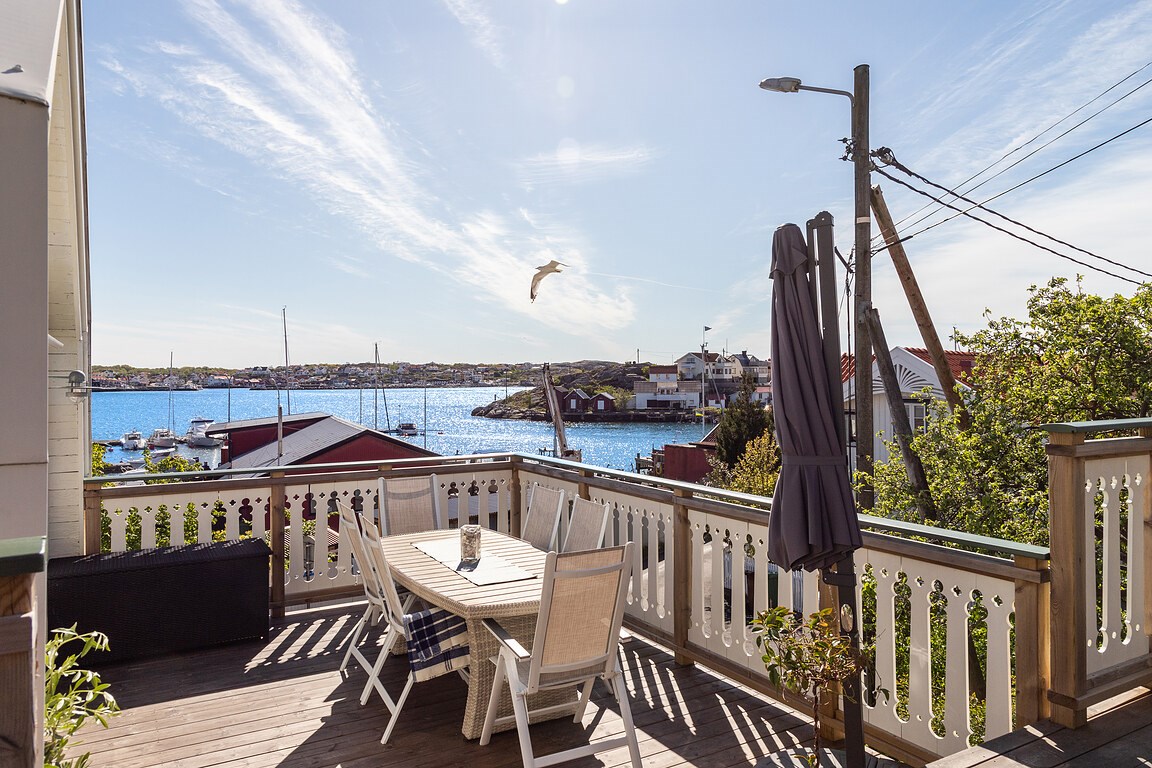 A spacious outdoor terrace is shown, featuring a dining table with several chairs and a large umbrella. The view overlooks a scenic harbor with boats, while vibrant greenery adds a touch of nature. Bright blue skies enhance the relaxing atmosphere.