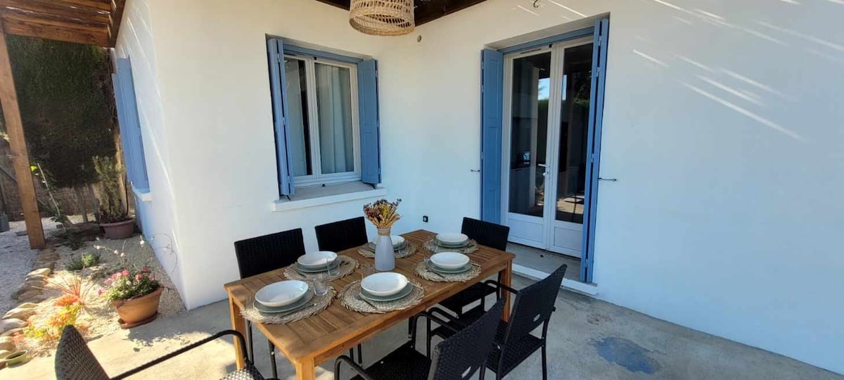 An outdoor dining area features a wooden table set for six, accompanied by black chairs. Two windows with blue shutters overlook the space, while a decorative light fixture hangs overhead, providing a cozy ambiance for al fresco meals.