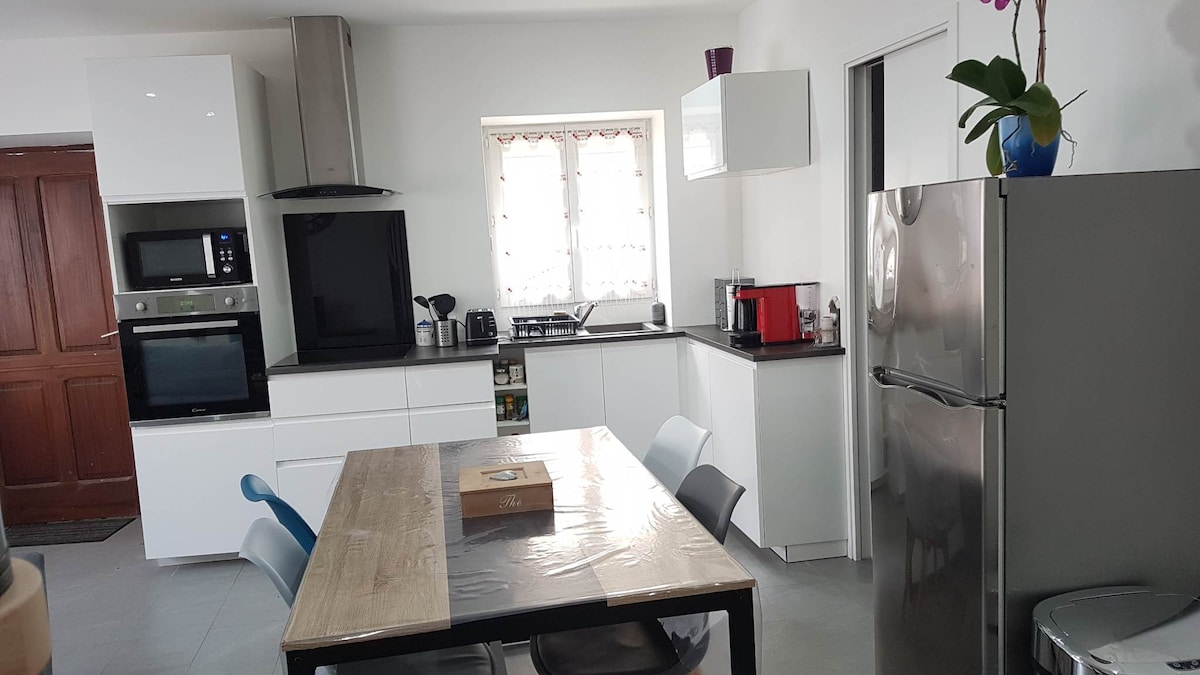 The kitchen features modern white cabinetry and grey countertops, complemented by stainless steel appliances, including an oven and refrigerator. A large dining table is surrounded by chairs, and natural light enters through the window, which is dressed with sheer curtains. A decorative plant adds a touch of greenery.