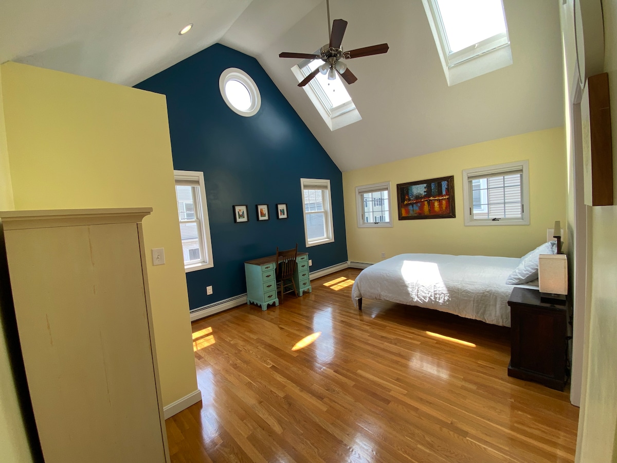 Deuxième chambre ensoleillée avec plafonds voûtés, lit Queen, espace de travail et salle de bain attenante.