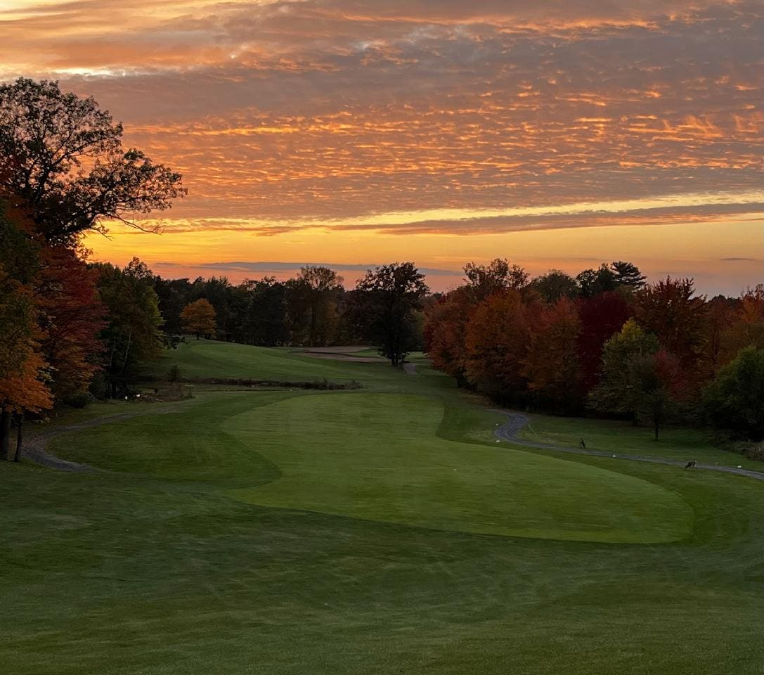 A lush green golf course spreads across the foreground, framed by vibrant trees showcasing autumn foliage. A soft orange and pink hue fills the sky as the sun sets, creating a serene atmosphere. The winding fairway leads into the distance, adding depth to the scene.