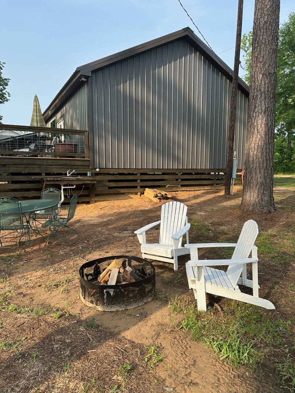 A fire pit rests on sandy ground, surrounded by two white Adirondack chairs. The exterior of the cabin, with vertical metal siding, is visible in the background, along with a wooden deck elevated above the ground. Tall trees provide a natural setting.