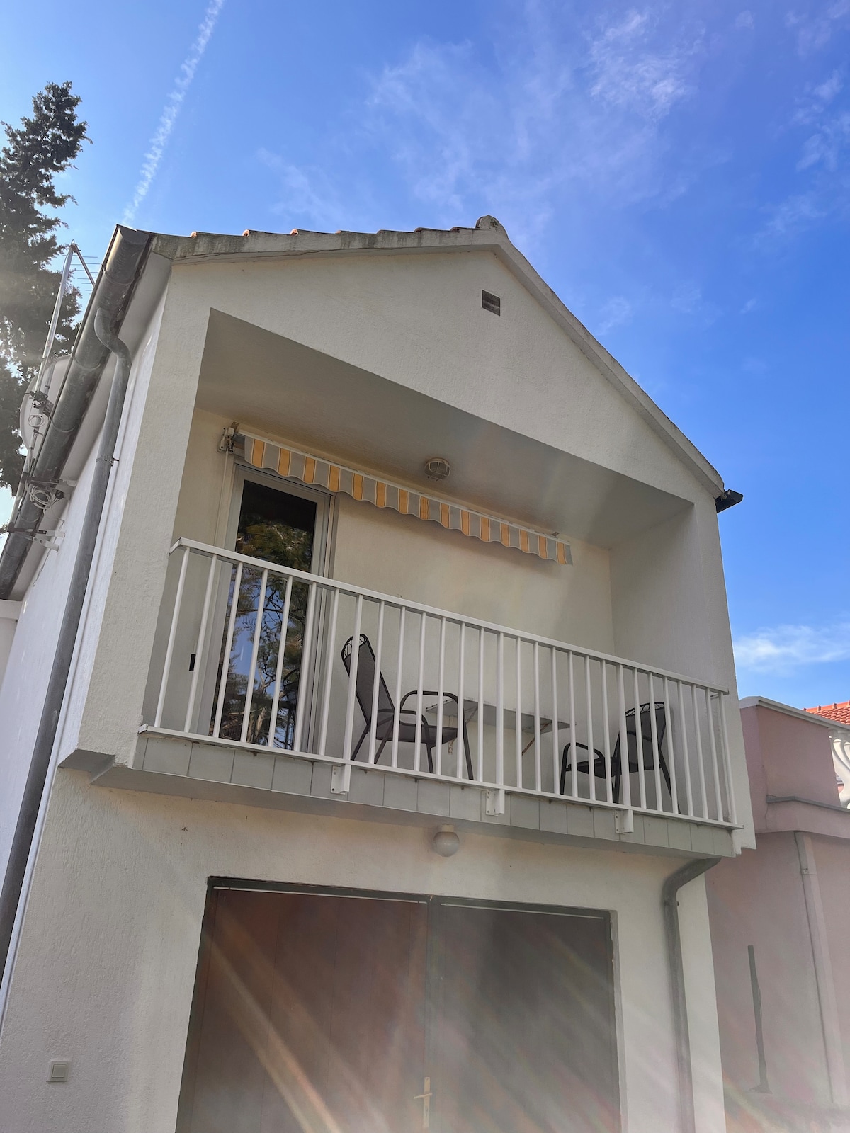 A white two-story building features a small, covered balcony with two streamlined chairs. The balcony railing is white, complementing the clear blue sky above. Sunlight beams create a bright atmosphere, enhancing the quietness of the surrounding area.