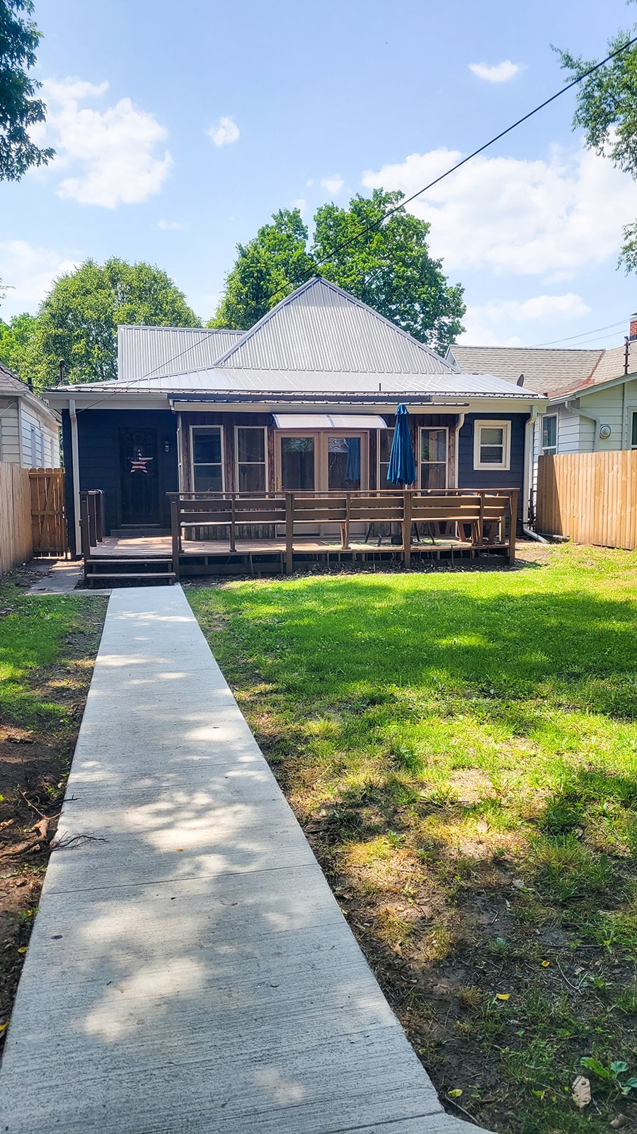 A craftsman-style house is featured with a welcoming front porch, complete with a blue umbrella. The spacious yard is surrounded by a wooden fence, and a concrete pathway leads to the entrance, which is framed by trees and greenery under a bright sky.