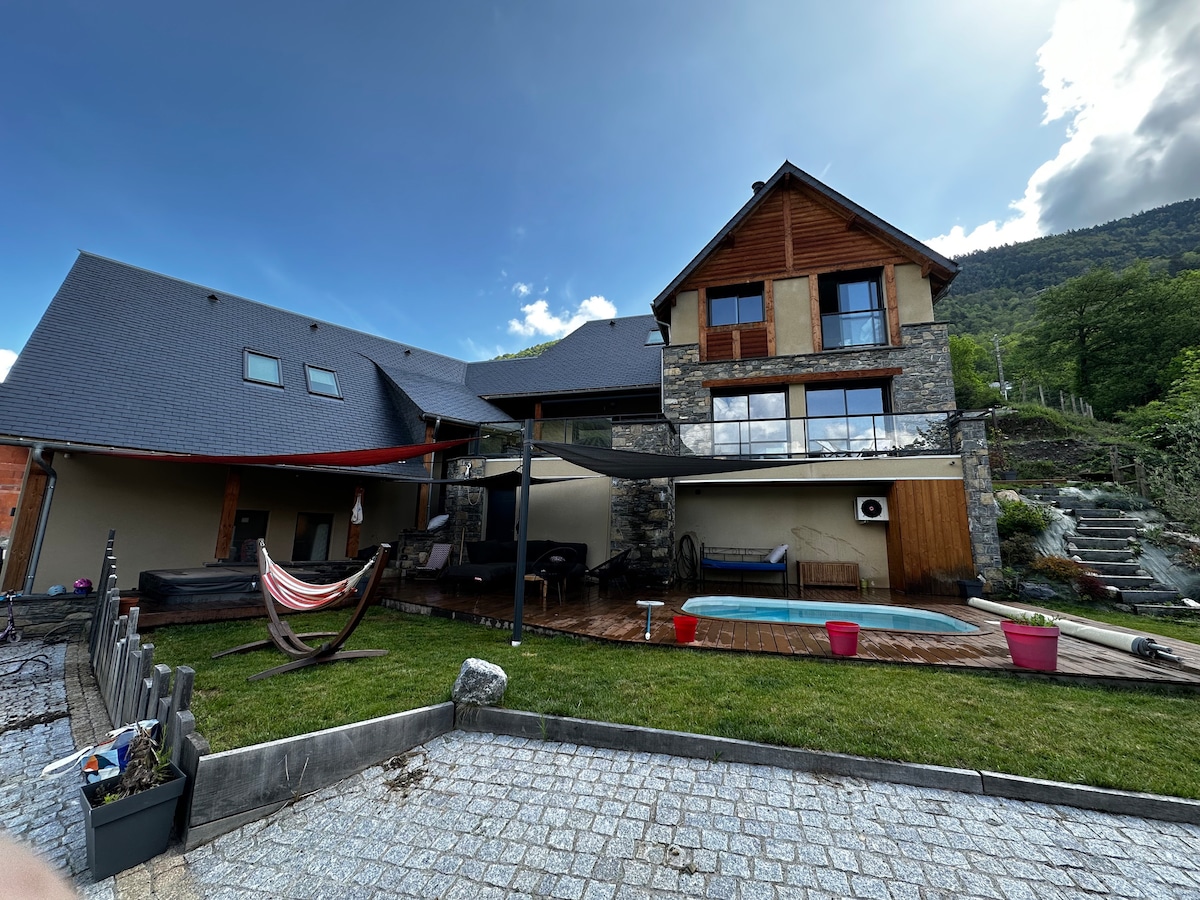 The exterior of the home is showcased, highlighting a spacious patio area with a swimming pool. Stone and wood elements combine with large glass windows, providing views of the surrounding greenery. A hammock is visible under a shaded area, contributing to a relaxed atmosphere.
