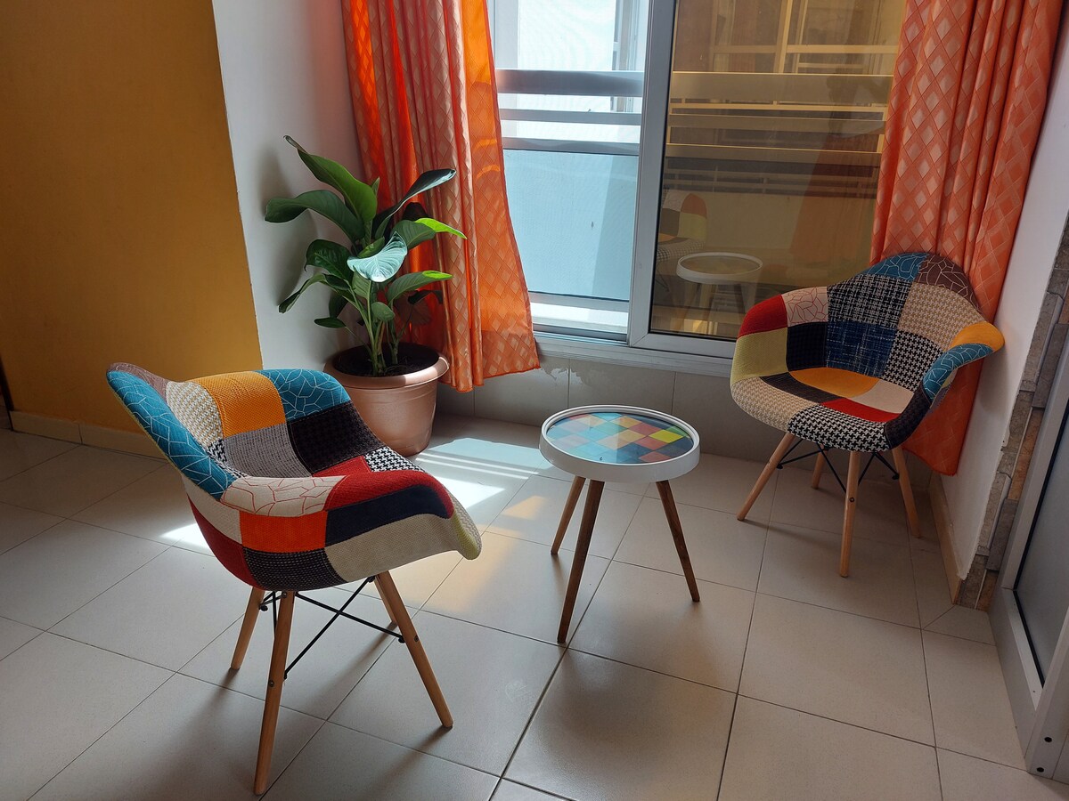 A bright corner of the living room features two comfortable armchairs with colorful fabric patterns, arranged around a small, round table. A potted plant adds a touch of greenery, and large windows are dressed with orange curtains, allowing natural light to fill the space.