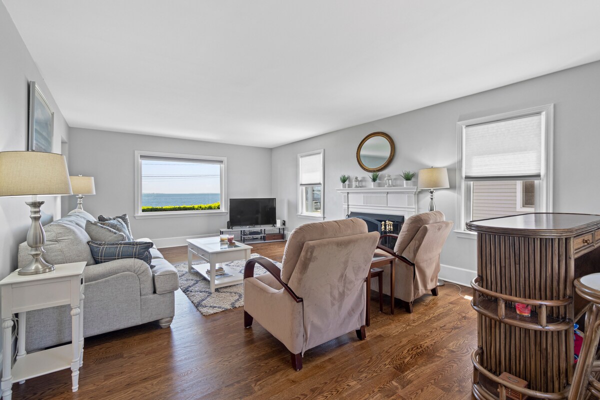 A bright living area features large windows offering views of Long Island Sound. Comfortable seating includes two armchairs and a sofa, complemented by a coffee table in the center. Light hardwood flooring enhances the space, and a fireplace adds a cozy touch.