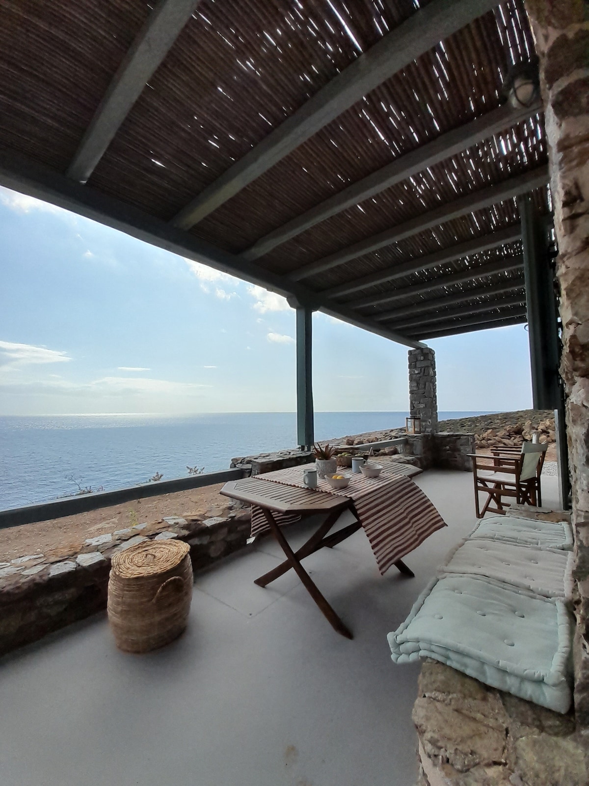 A shaded terrace offers a view of the sea, with a wooden table set for dining. Stone walls frame the space, complemented by woven details and a natural fiber basket. Light clouds pass overhead, enhancing the tranquil ambiance.