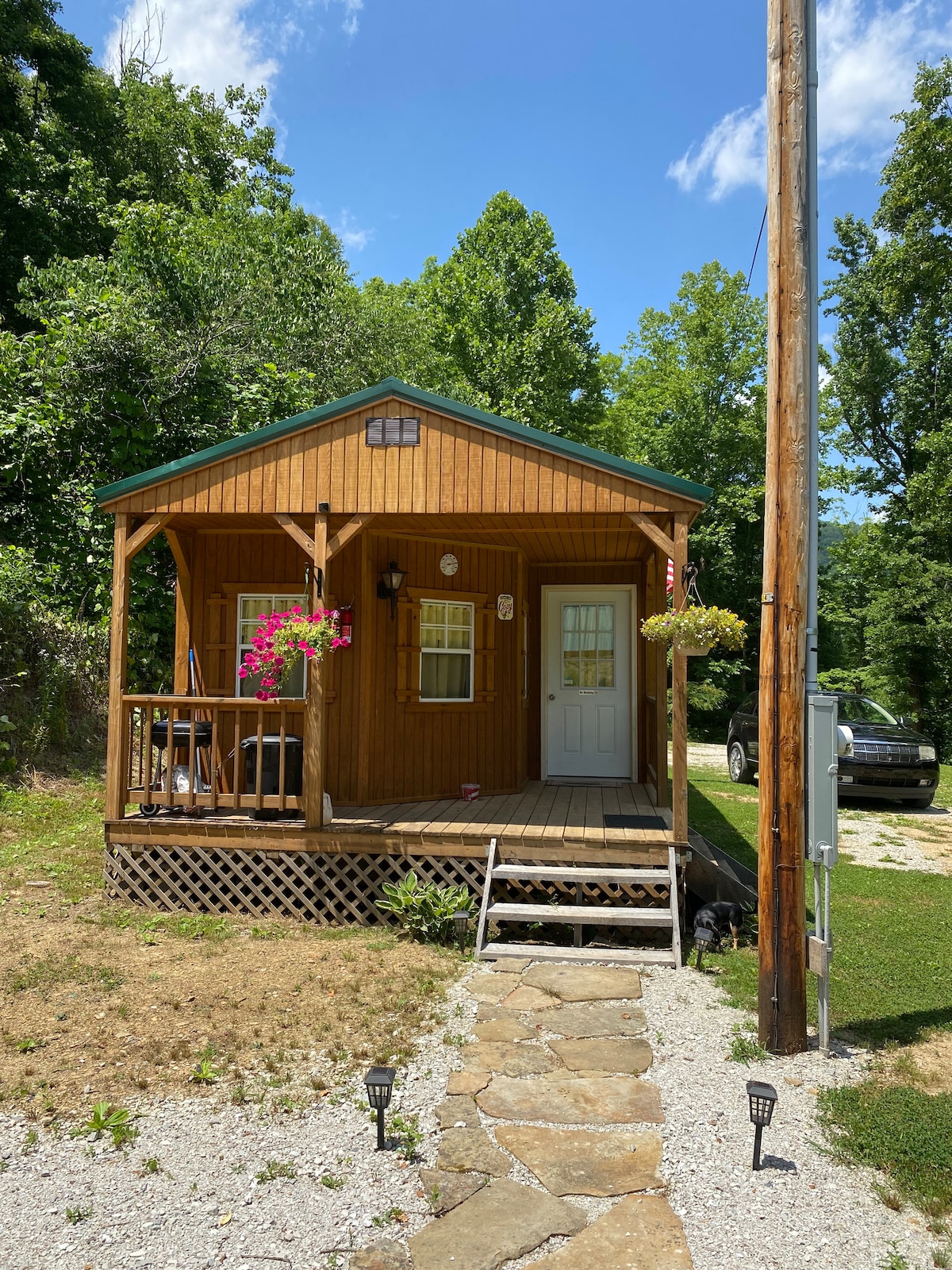A charming wooden cabin is showcased with a welcoming porch adorned with hanging flower planters. The entrance is framed by greenery and a stone pathway leads up to the steps. A driveway is visible beside the cabin, surrounded by a grassy area.