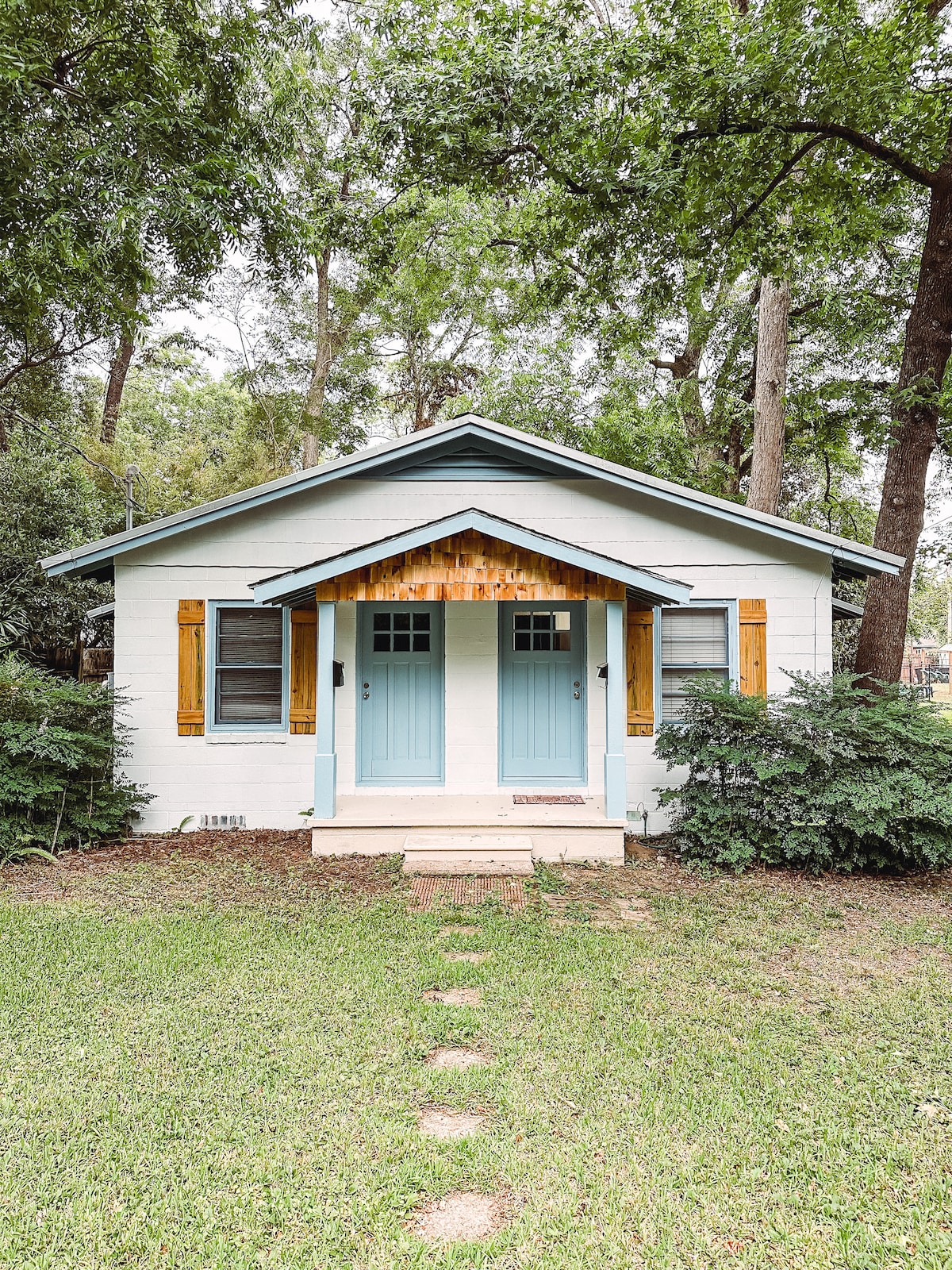 A charming duplex is framed by lush greenery, featuring a well-maintained lawn and stone pathway. The front entrance is highlighted by two blue doors with wooden accents, and rustic shutters complement the white exterior of the home.