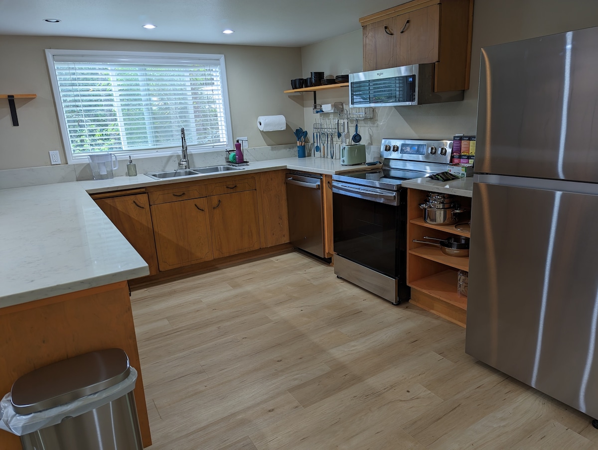 The kitchen features wooden cabinetry, modern stainless-steel appliances, and a spacious countertop. Natural light floods the area through a large window. Utensils and kitchenware are neatly organized on shelves, and a trash bin is positioned discreetly in the corner.
