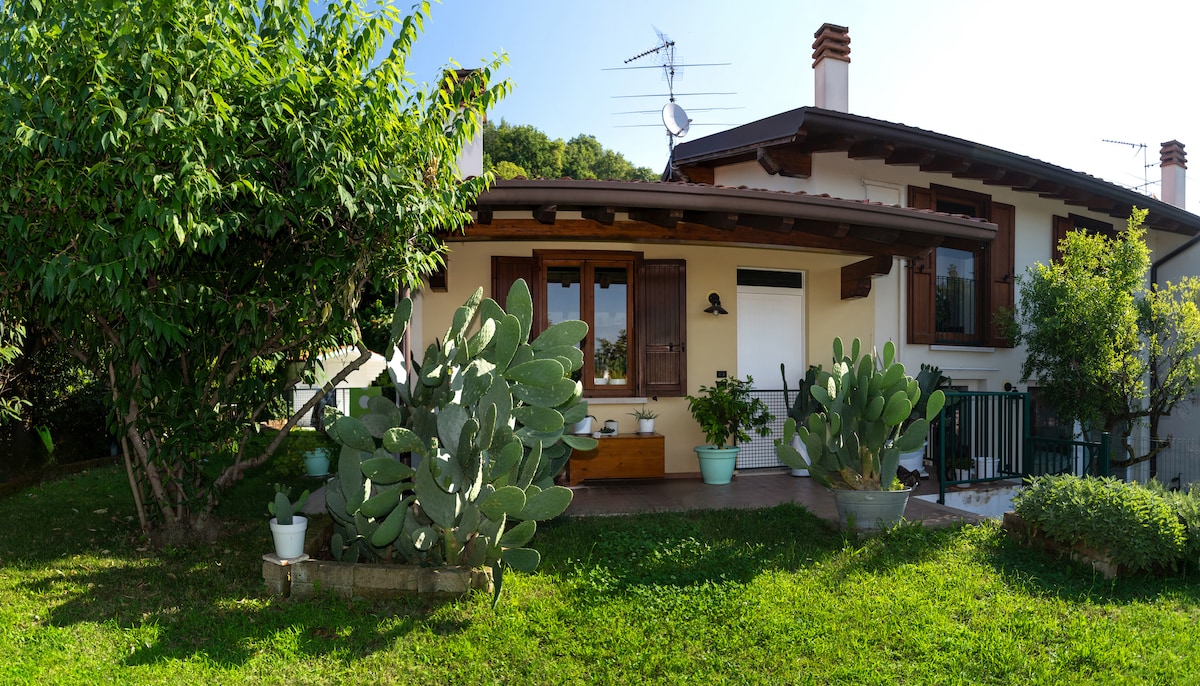 The image showcases the exterior of a charming building surrounded by greenery. A cozy porch area is visible, adorned with potted plants, including cacti. The structure features inviting wooden details and large windows that allow natural light to fill the space.
