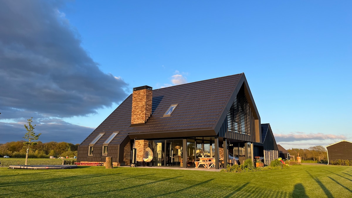 A contemporary-style house with a distinctive triangular roof is surrounded by lush green grass. Large glass doors and windows offer expansive views of the landscape, while a stone chimney adds an earthy element to the modern design.