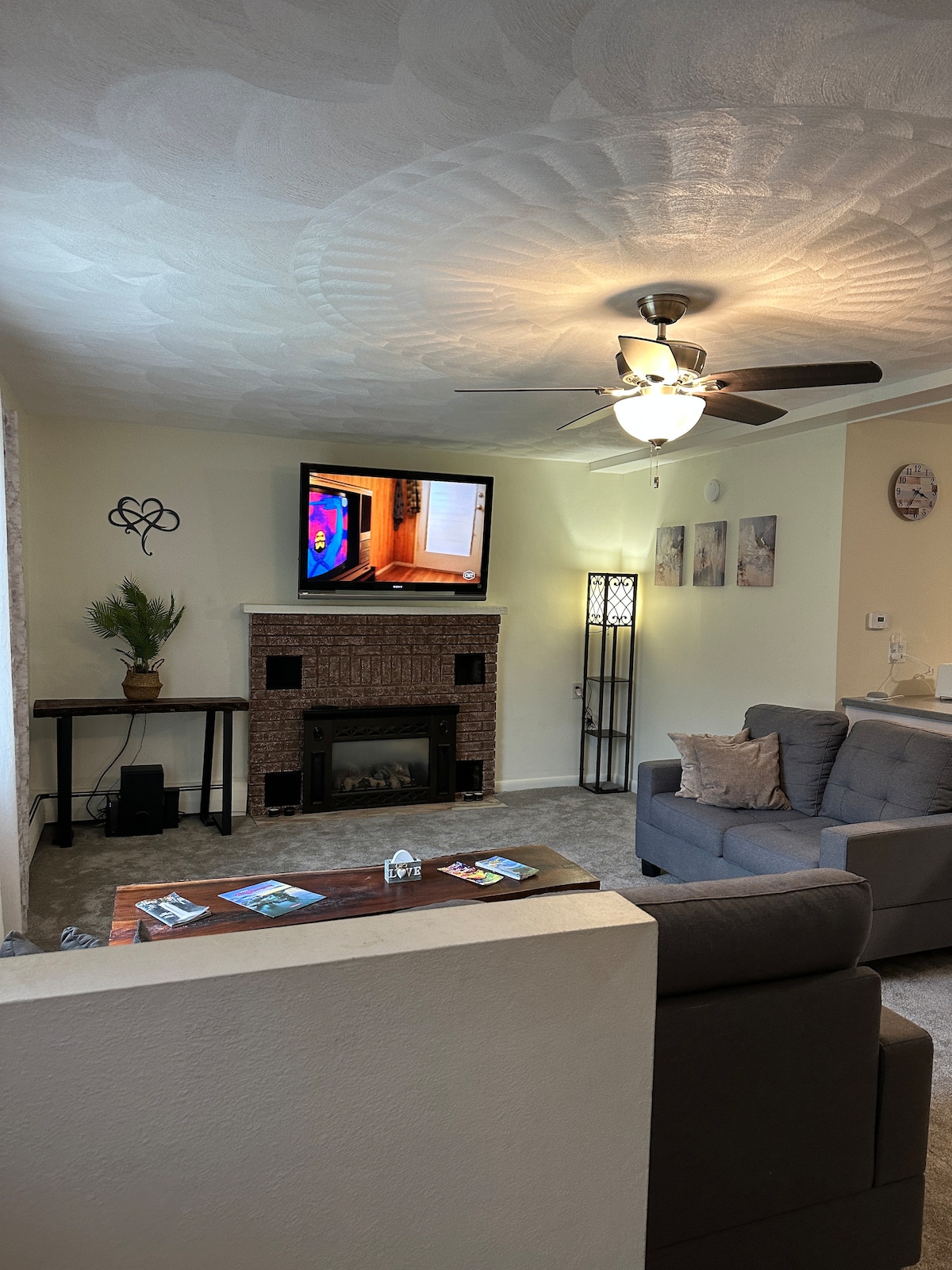 A cozy living room is presented, featuring a television mounted above a decorative fireplace. A ceiling fan is installed to enhance air circulation. A comfortable gray sofa is positioned in the seating area, alongside a small table and a decorative plant in the corner.