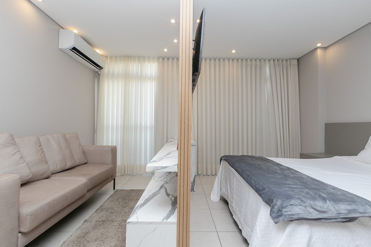 A modern living space is divided by a wooden partition, showcasing a comfortable sofa and a marble table. The sleeping area features a spacious bed with a soft gray blanket, illuminated by natural light filtering through sheer curtains.