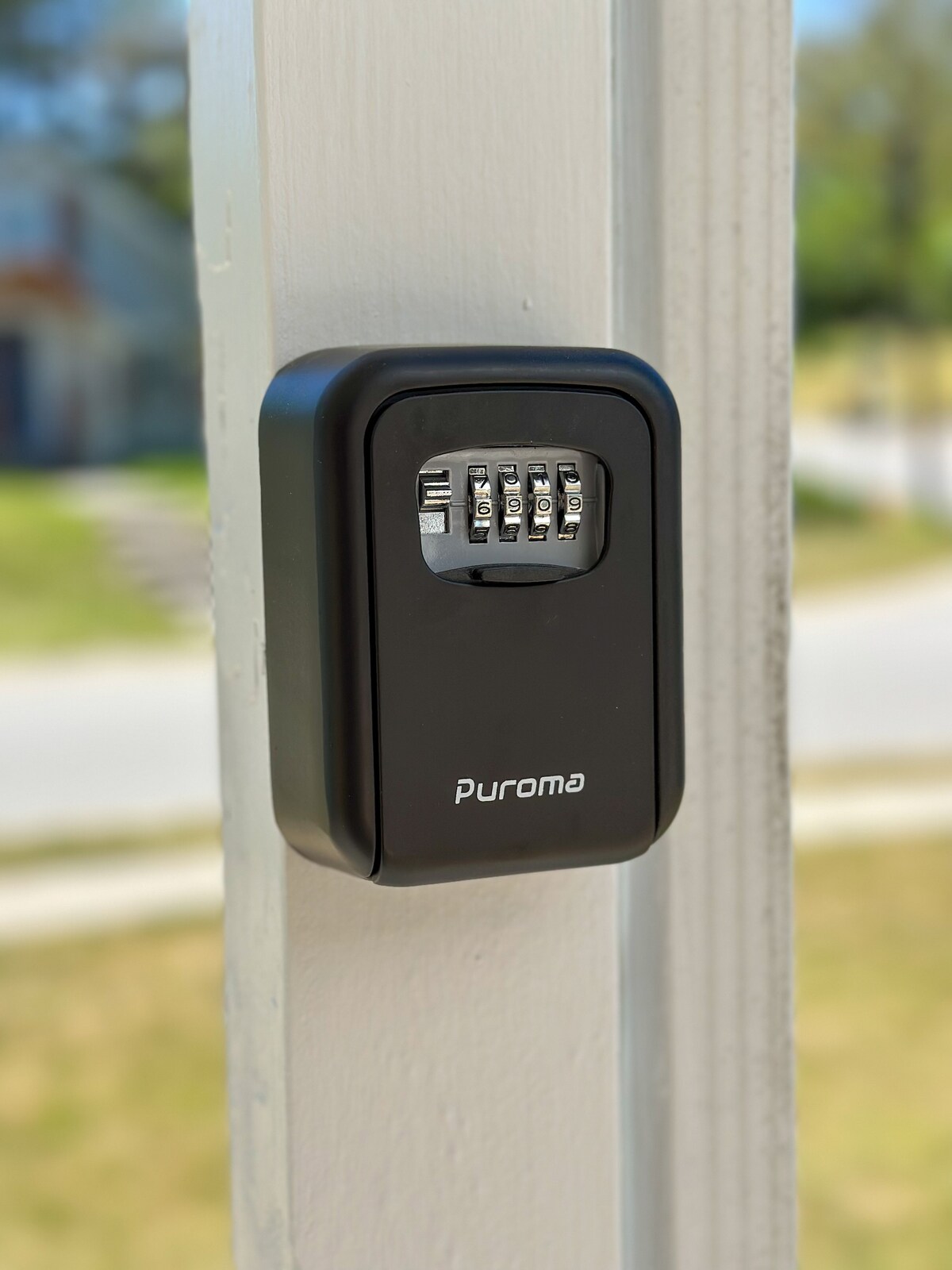 A secure combination lockbox is mounted on a light-colored wall, designed for key storage. The lock features a four-digit combination mechanism. A blurred view of a grassy area and paved street is visible in the background.