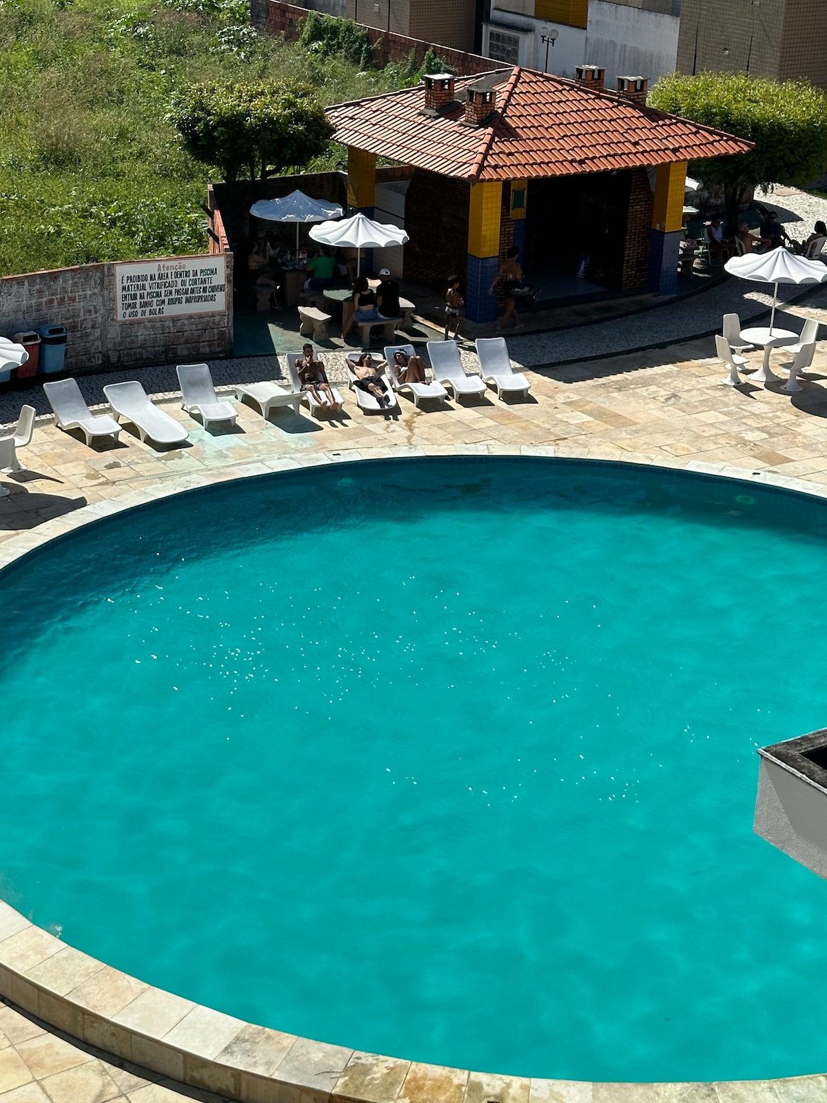 A large swimming pool is featured, surrounded by lounge chairs and shaded by umbrellas. The pool's clear turquoise water sparkles under the sun, while a nearby building offers additional seating and shade, inviting relaxation and leisure.