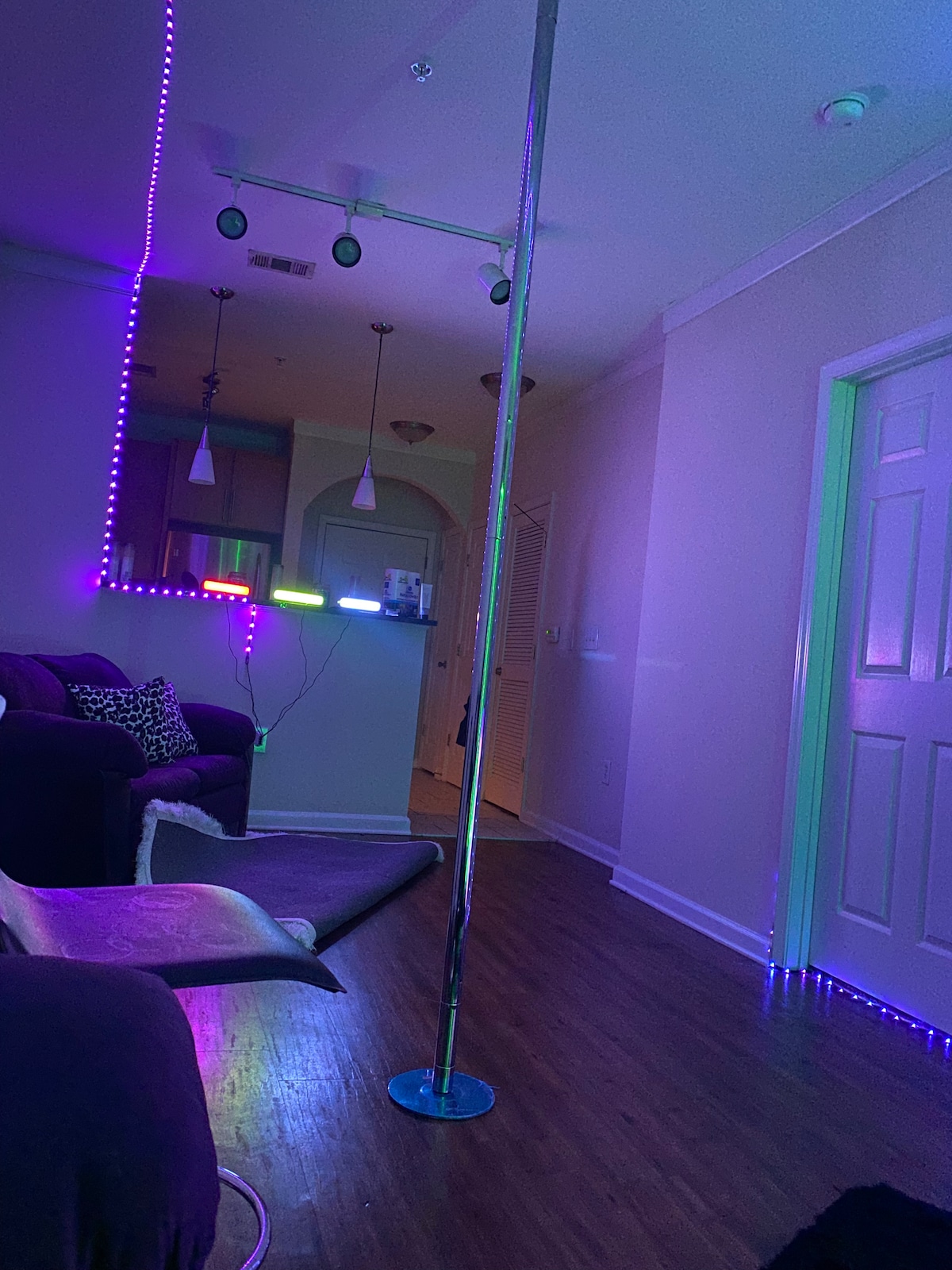 A living room space features a metallic stripper pole illuminated by colorful LED lights. A plush couch is located nearby, with a pet bed visible on the floor. The adjacent kitchen is discernible through a doorway, showcasing pendant lighting and modern appliances.