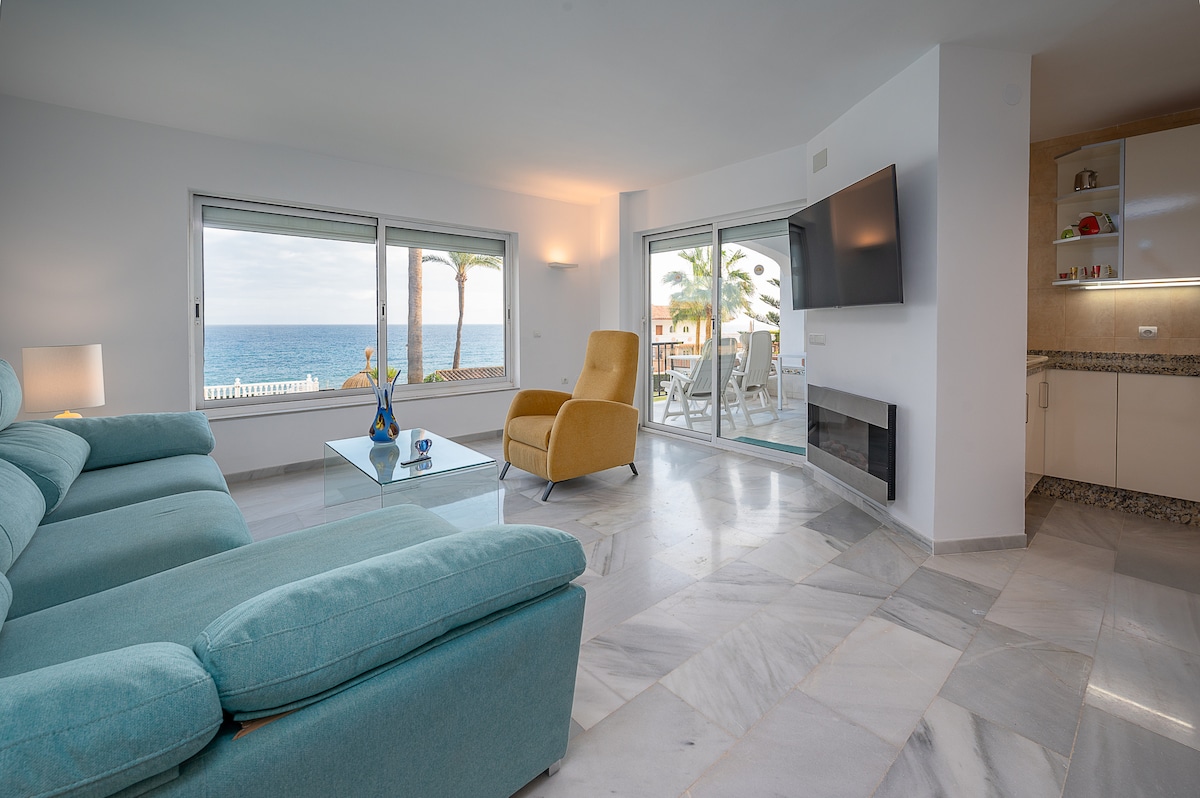 A light-filled living area is furnished with a comfortable turquoise sofa and an accent chair. A glass coffee table sits in front. Large windows display views of palm trees and the ocean. A wall-mounted TV and a modern fireplace enhance the space.