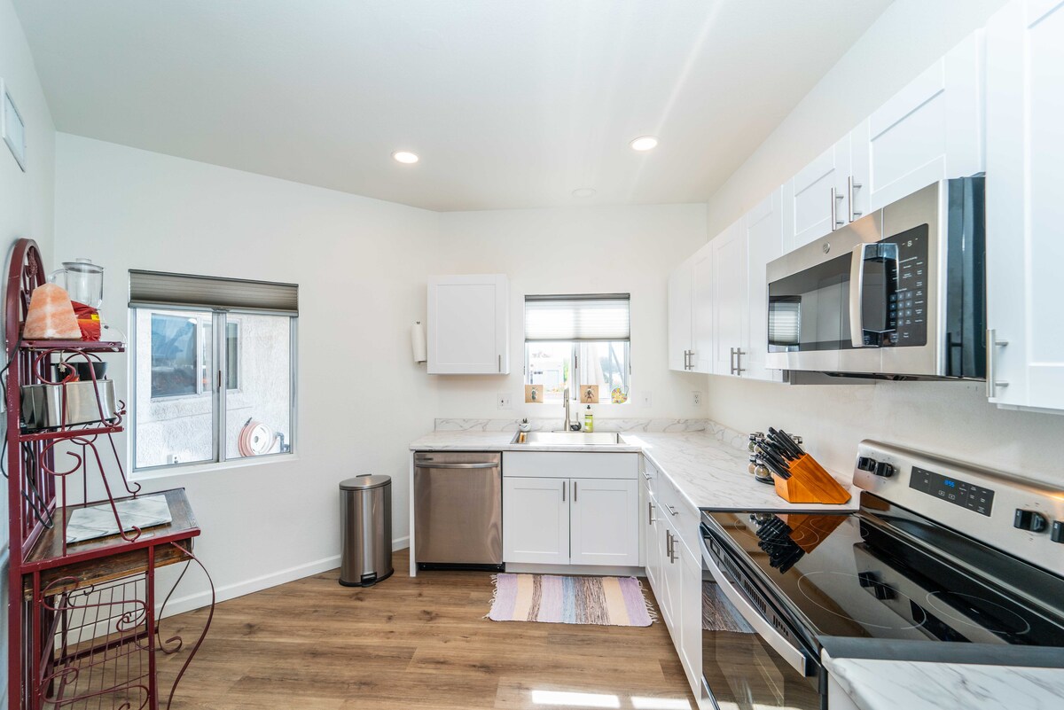 A bright kitchen is equipped with modern stainless steel appliances, including a refrigerator, microwave, and oven. White cabinetry contrasts with the marble countertops. Natural light enters through a window, enhancing the spacious feel. A wood floor and ample storage complete the inviting design.