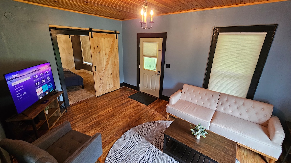 A living area displays a light-colored sofa and a gray armchair, positioned around a dark wooden coffee table. A large television is mounted on the wall, and sliding barn doors lead to a bedroom. Natural light filters through the window, illuminating the wooden floor.