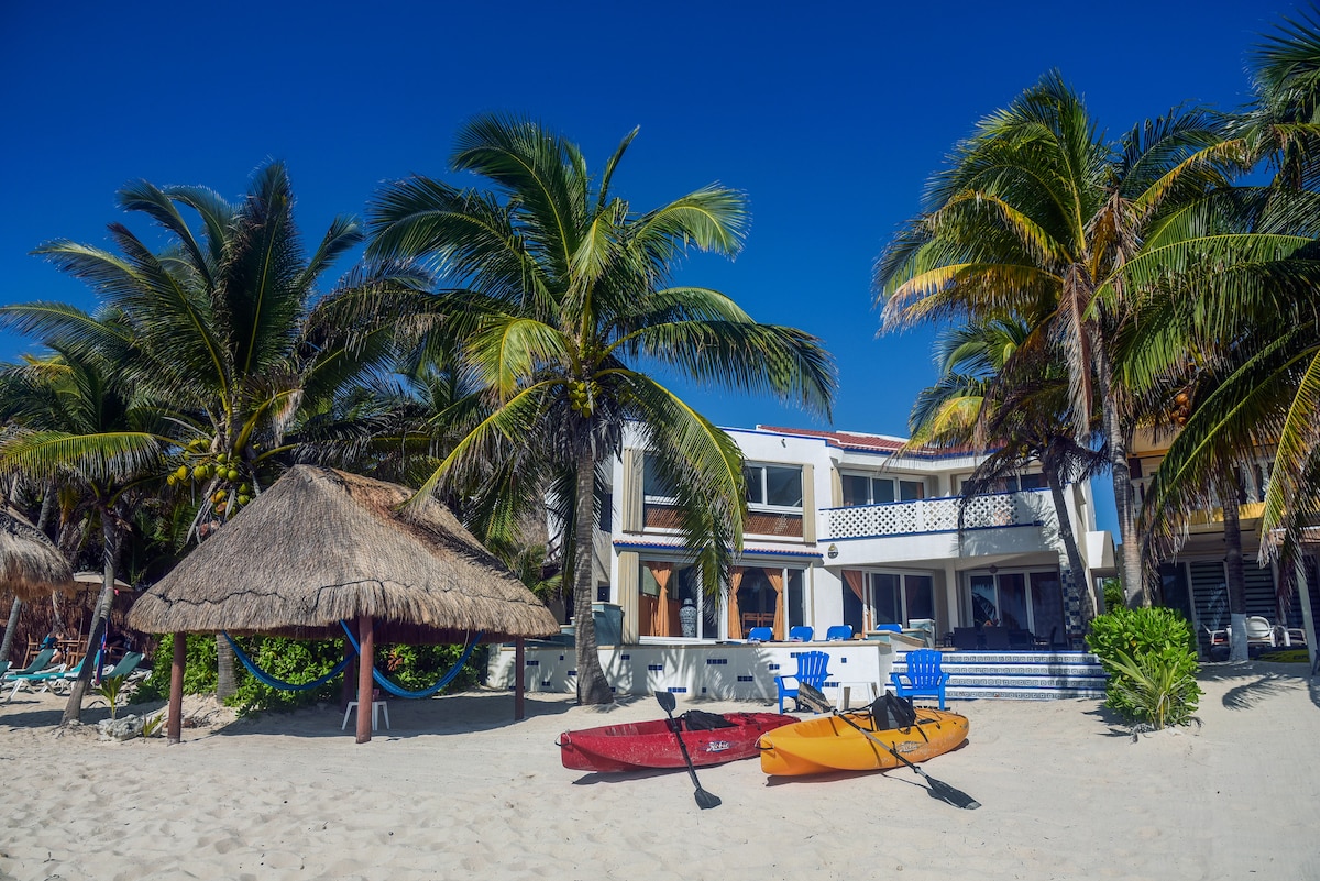 A beachfront property is framed by tall palms, with colorful kayaks resting on the soft sand. A thatched-roof palapa provides shade, while white lounge chairs surround a table near the villa's entrance, smoothly transitioning from the outdoor space to the house.